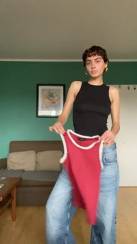 Ung person med kort hår, iført en sort ærmeløs top og jeans, der holder en rød ærmeløs skjorte indendørs ved en sofa og en grøn væg.
