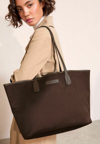 Brązowa torba typu tote z gładkiego materiału, kontrastującymi szarymi uchwytami i metką z napisem "przyjaciele tacy jak oni". Strukturalny kształt z otwartą górą.