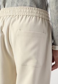 Beige Jogginghose mit elastischem Bund, ausgestattet mit einer einzigen aufgesetzten Tasche auf der Rückseite und einer strukturierten Stoffoberfläche.