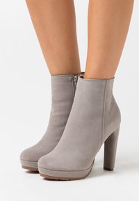 Botines de ante gris claro con cremallera lateral y tacones altos, llevados sobre piernas descubiertas contra un fondo blanco.
