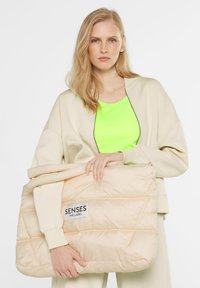 Cremfarbener Pufferbeutel mit einem gesteppten Design, Tragriemen und einem eingenähten Etikett; getragen zu einem leuchtend grünen Oberteil und einer neutralen Jacke.