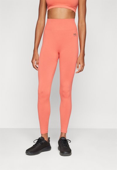 Korallenfarbene High-Waist-Leggings aus elastischem Stoff, mit einer taillierten Passform und dezentem Branding an der Seite, kombiniert mit schwarzen Sportschuhen.