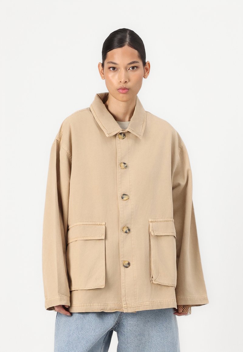 Beige oversized jacket made of cotton canvas, featuring a collar, five buttons, and two front pockets. Details include stitching and a relaxed fit.
