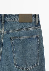 Jeansy denimowe w średnim odcieniu niebieskiego z teksturowaną powierzchnią. Posiadają skórzaną metkę z tłoczonym tekstem na tylnej części pasa oraz tylne kieszenie.