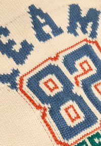 Gros plan sur un tissu tricoté avec le grand chiffre « 86 » en contour bleu et orange, et un texte bleu partiel sur fond crème.