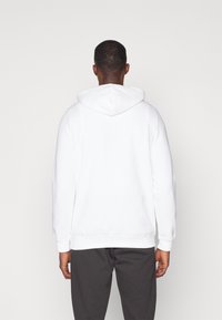 Hvid hættetrøje med snor i hætten, ribbede manchetter og kant, parret med mørkegrå sweatpants. Stoffet ser blødt og utextureret ud.