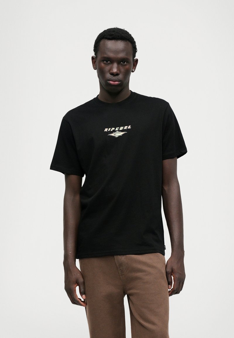 T-shirt de algodão preto com um logótipo central "Rip Curl" em branco e prata sob um gráfico de uma onda; combinado com calças castanhas.