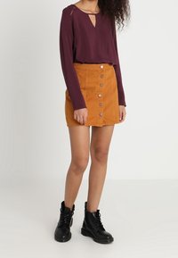 Blusa bordeaux a maniche lunghe con scollatura a chiave abbinata a una gonna in velluto a coste arancione con bottoni frontali e stivaletti neri alla caviglia.