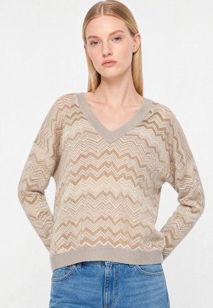 Junge Frau mit glatten blonden Haaren, die einen beige-weißen V-Ausschnitt-Pullover mit Zickzackmuster und blaue Jeans trägt, steht mit den Händen hinter dem Rücken.