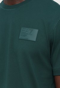 Zaļš kokvilnas t-krekls ar kvadrātveida gumijotu logotipa uzlīmi kreisajā krūtīs. Audums ir ar gludu tekstūru un apļveida izgriezumu kaklā.