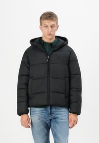 Sort pufferjakke med hætte, lynlås og vandret syninger. Båret over en grøn skjorte og parret med lyseblå jeans.