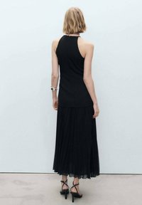 Čierny top s halter krkom doplnený o splývavú čiernu maxi sukňu. Materiál vyzerá ľahký s detailami v podobe plisovania. Čierne sandále na opätku.