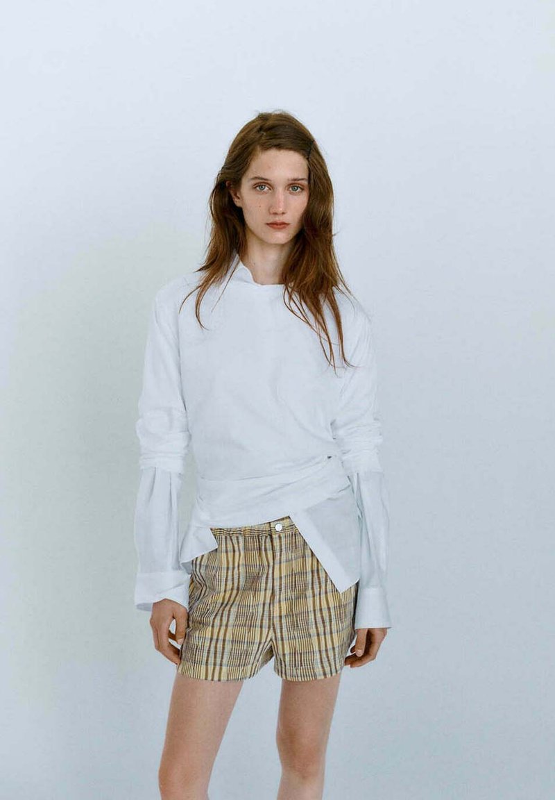 Jeune femme aux longs cheveux bruns portant une chemise blanche à manches longues et un short jaune à carreaux, debout devant un fond clair uni.