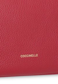Červená kožená peněženka s texturovaným povrchem a embosovaným zlatým logem "Coccinelle". Jednoduchý design bez dalšího hardwaru nebo vzorů.