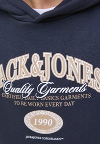 Marineblauer Hoodie mit auffälligem, cremefarbenem Text. Das Design enthält "JACK&JONES", "Qualitätsbekleidung" und ein Emblem von 1990 am unteren Rand. Weicher Stoff.