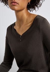 Haut à manches longues, marron foncé, avec un décolleté en V ornée de deux boutons noirs, confectionné dans un tissu doux et lisse, affichant une silhouette ajustée.