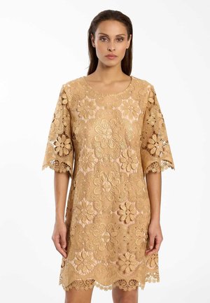 Femme portant une robe en dentelle beige longueur genoux avec des motifs floraux et un ourlet festonné, debout devant un fond blanc.