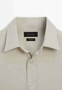 Jasno beżowa koszula z bawełny i elastanu, z klasycznym kołnierzykiem, zapięciem na guziki z przodu oraz czarną metką ze złotym napisem. Gładka tekstura.