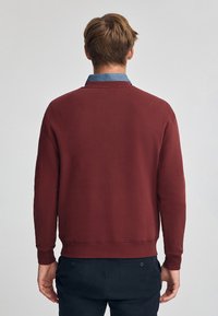 Burgundy sweatshirt med långa ärmar, rund halsringning och ribbade muddar. Mjuk textur, minimalistisk design, bärs över en blå skjorta med krage.