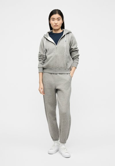 Νεαρή γυναίκα που φοράει γκρι ζακέτα με φερμουάρ, ταιριαστό παντελόνι φόρμας, μπλε μπλούζα, λευκές κάλτσες και λευκά αθλητικά παπούτσια, με φόντο ένα απλό τοπίο.