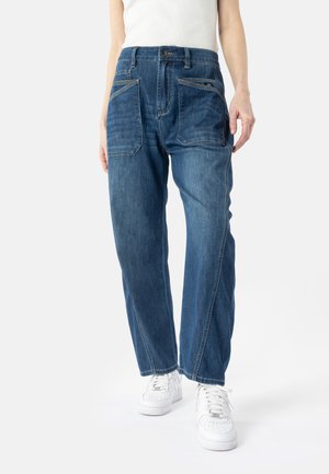 Kvinde iført løstsiddende blå jeans med store forlommer, kombineret med hvide sneakers og en hvid ærmeløs top, stående på en hvid baggrund.