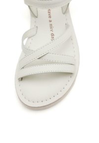 Sandalia de cuero blanco con tiras entrelazadas y detalles de costura. Diseño de punta abierta con una suela plana y suave. El texto interno dice "que tengas un buen día".