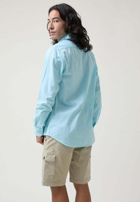Camisa de manga larga azul claro con puños abotonados, combinada con pantalones cortos cargo beige claro que tienen bolsillos laterales y un dobladillo justo por encima de la rodilla.