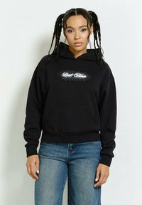 Czarna bluza z kapturem z przednią kieszenią, z białym wzorem tekstowym i akcentami w kropki. W połączeniu z niebieskimi dżinsami, które prezentują swobodny krój.