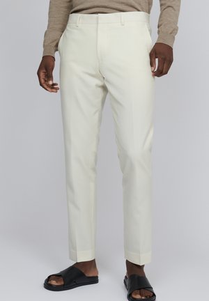 Pantalons beige clair avec une coupe slim, dotés d'un devant plat et d'un pli subtil. Tissu lisse avec un léger éclat, associés à des sandales noires à glissière.