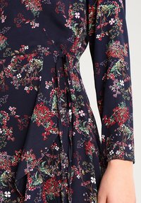 Blumenkleid mit Marineblauem Hintergrund, bunten Blumenmustern in Rot, Rosa und Grün, mit gebundenem Taillenbund und langen Ärmeln.