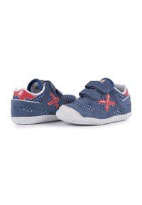 Des chaussures en daim bleu avec des pois blancs, des accents rouges et une semelle texturée blanche. Elles sont dotées de brides en Velcro pour la fermeture.