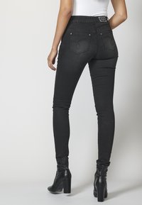 Kvinna iklädd svarta skinny jeans och svarta klackade ankboots, står vänd bort mot en plain vit bakgrund.