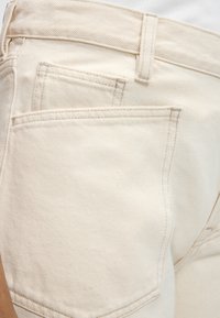 Κοντινή λήψη εκρού τζιν που δείχνει πίσω τσέπη, θηλιά για ζώνη και λεπτομέρειες ραφής.