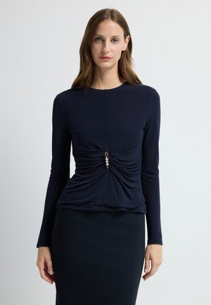 Vrouw in een aansluitend marineblauw longsleeve top met geplooide details en een decoratieve sluiting op de taille, gecombineerd met een bijpassende rok.