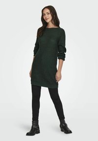 Abito in maglia di colore verde scuro con scollatura rotonda e maniche lunghe arrotolate. Abbinato a jeans skinny neri e stivaletti.