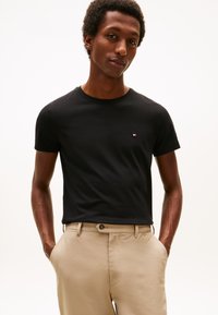 Črna bombažna majica z okroglim izrezom in kratkimi rokavi, z majhnim vezenim logotipom na prsih, v kombinaciji z bež hlačami.