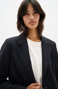 Black blazer with notched lapels, single button closure, and a smooth texture, worn over a white shirt. Model has shoulder-length hair.
