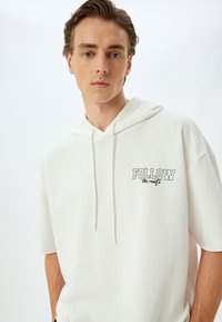 Witte hoodie met een voorzak, korte mouwen en een capuchon met trekkoord. Heeft de tekst "FOLLOW the route" in het zwart op de borst gedrukt. Gladde textuur.