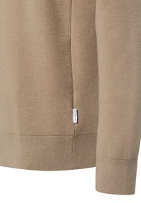 Nahaufnahme des Saums und Ärmelsaums eines beigen Strickpullovers mit kleinem weißen Markenschild an der Seitennaht.