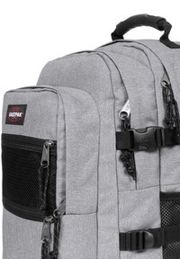 Mochila de tela gris con múltiples compartimentos, acentos de malla negra, cremalleras y un cierre negro. Presenta un asa superior y un parche con el logo.