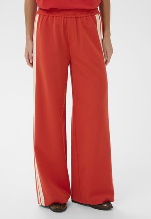 Partie inférieure du corps portant un pantalon rouge à jambes larges avec des bandes blanches sur les côtés et des chaussures marron sur un fond blanc uni.