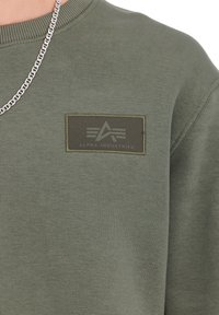 Felpa color verde oliva con superficie testurizzata. Presenta una patch rettangolare con il logo "ALPHA INDUSTRIES" e un dettaglio collana in catena argentata.