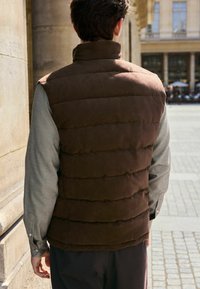 Brun puffer vest med høj krave, quilted tekstur og en grå langærmet skjorte nedenunder. Baggrunden viser en stenmur og udendørs siddepladser.