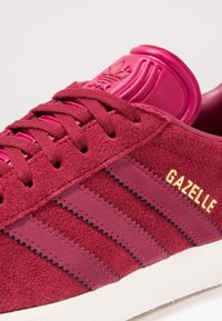 Röda mockasneakers med tre mörkare röda ränder, en texturerad vinröd hälrem och guld "GAZELLE"-logotyp på sidan.