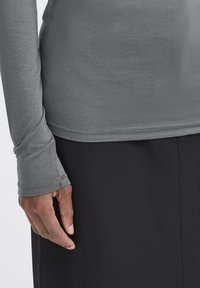 Mano appoggiata di lato, con una maglietta grigia a maniche lunghe con foro per il pollice e pantaloni neri, mostrata dalla vita alla mano.