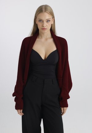 Jeune femme aux cheveux blonds raides porte un haut noir et un pantalon avec un cardigan tricoté ample rouge foncé sur un fond uni.