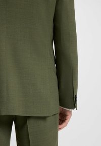 Olivgrüner Blazer mit strukturiertem Finish, ausgestattet mit einem Schlitz hinten und Knopfdetails an den Ärmeln. Strukturierte Passform und maßgeschneiderte Gestaltung.