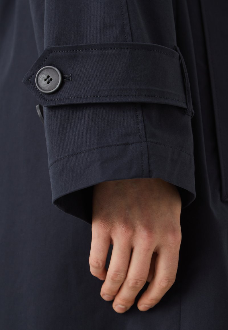 Polsino di un trench coat blu navy con chiusura a bottone, cuciture distintive e texture liscia, che mette in mostra una vestibilità sartoriale e un design strutturato.