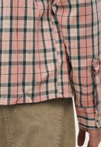 Gros plan d'une personne portant une chemise à carreaux rose, noir, blanc et turquoise, ainsi qu'un pantalon beige, montrant la manche de la chemise et le côté du torse.