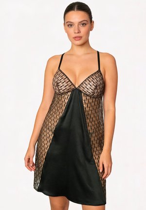 Donna che indossa un vestito sottoveste in raso nero e pizzo con spalline sottili, in piedi davanti a uno sfondo chiaro e uniforme.